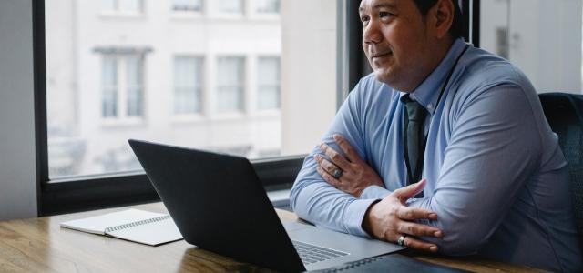 Professional-looking man sitting in an office. There is a laptop and a stack of files in front of him.